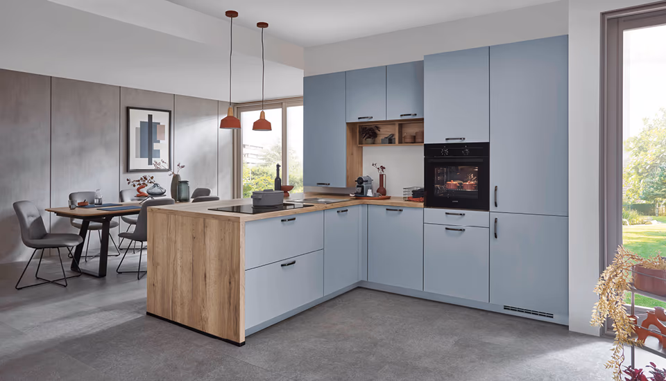 Stilvolle L-Küche in Hellblau mit Holzarbeitsplatte, Kochinsel und angrenzendem Essplatz in offenem Raum, ideal für modernes Wohnen mit harmonischem Farbkonzept.